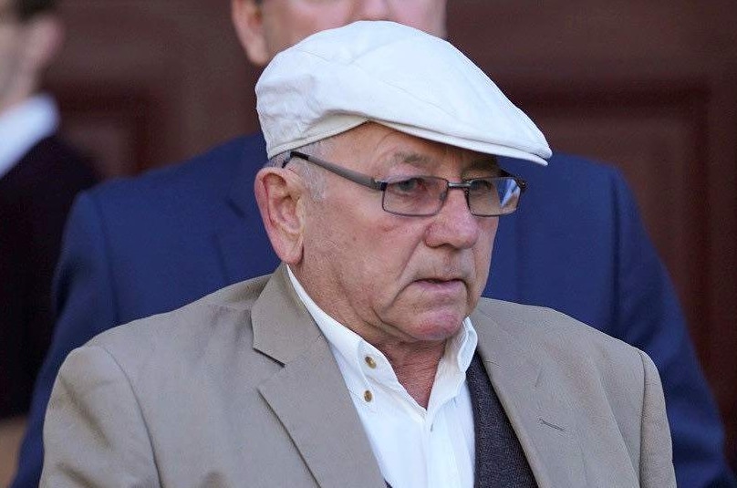 A older man with a grey jacket and white flat cap in front of court.