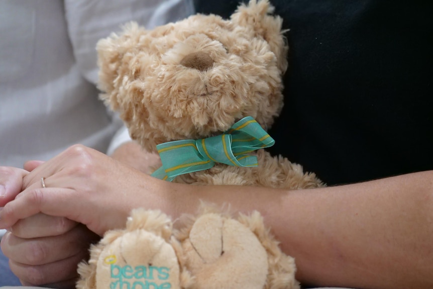 A brown teddy bear with a blue ribbon sits in a womans lap. 