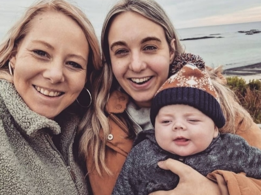 A smiling couple with their baby all rugged up with the ocean in the background.