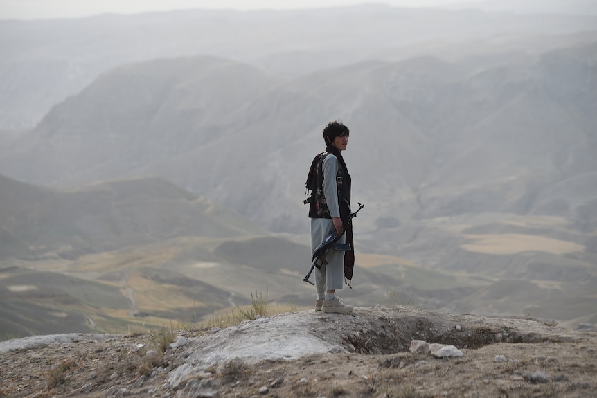 A young man stands on a cliff edge looking out over a wide valley. 