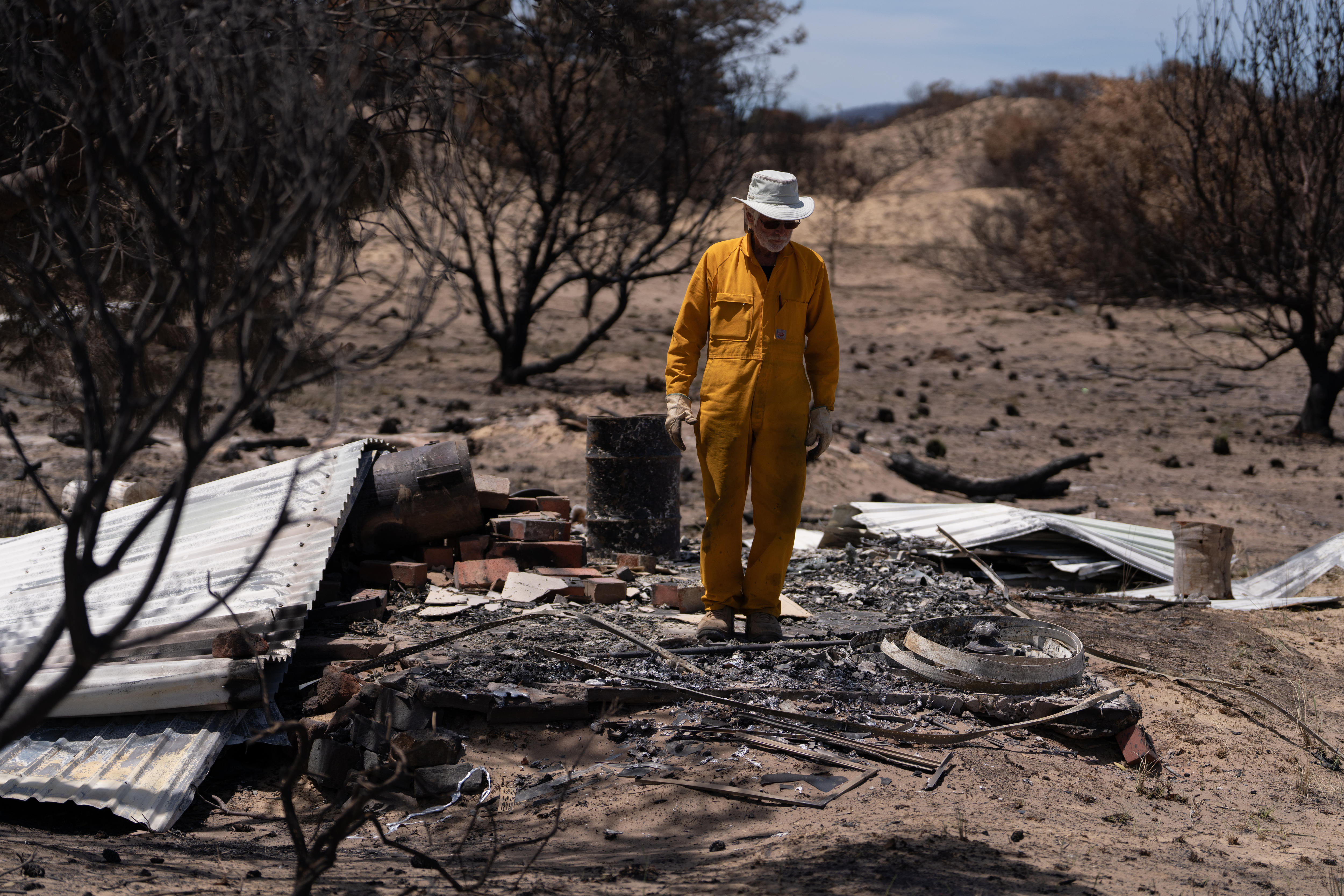 Un hombre vestido con un mono naranja y un sombrero de ala ancha se encuentra entre los escombros de su sauna después de un incendio forestal.
