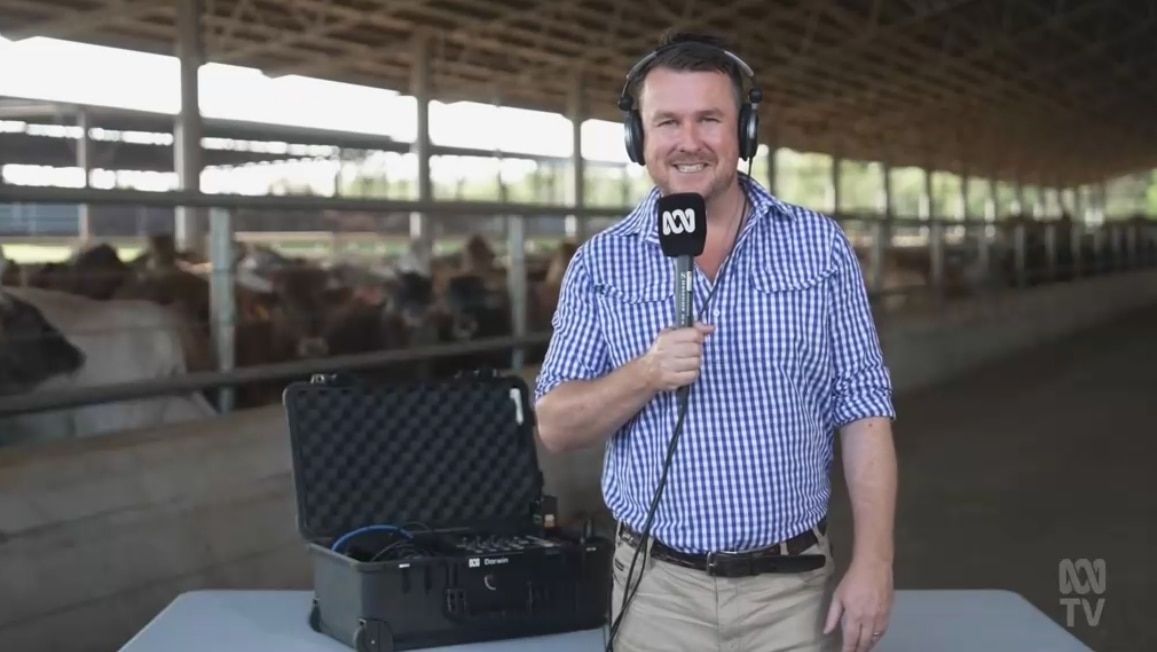 A man in headphones holding a microphone smiles to camera, cows behind him 