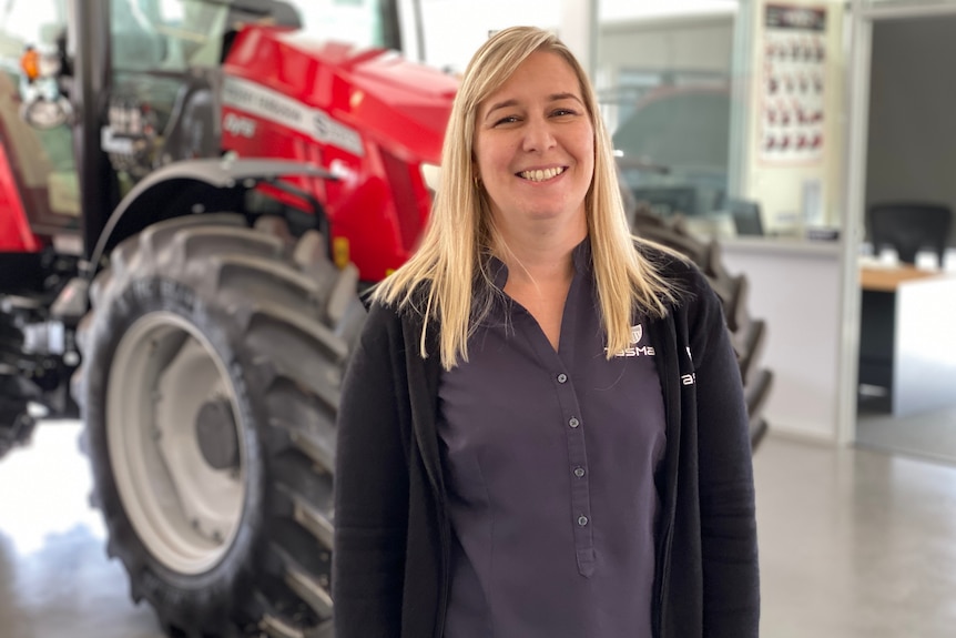 a blonde woman stands in a showroom-like space with a red tractor in the background