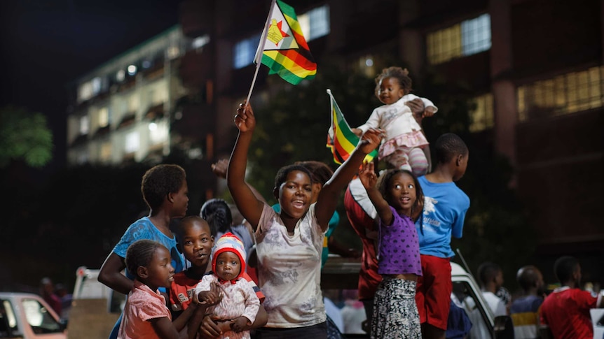 Zimbabweans stand on the back of a ute celebrate waving flags, smiling