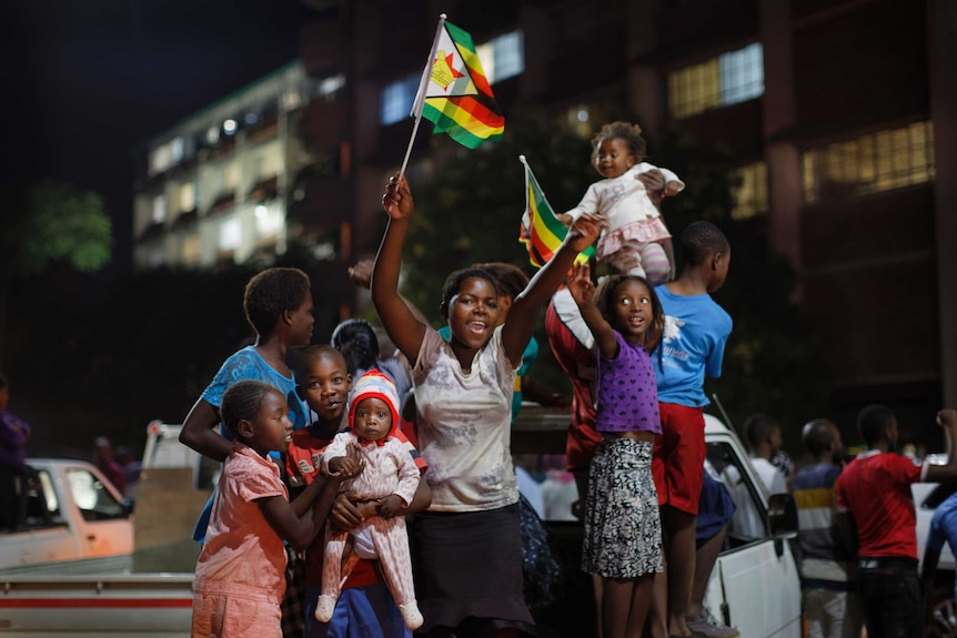 Zimbabweans stand on the back of a ute celebrate waving flags, smiling