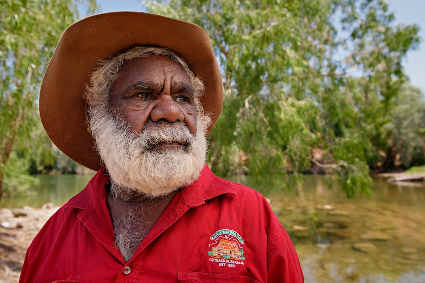 Un homme à l'air sérieux avec un chapeau et une chemise rouge regardant sur le côté, avec une rivière et une brousse en arrière-plan.
