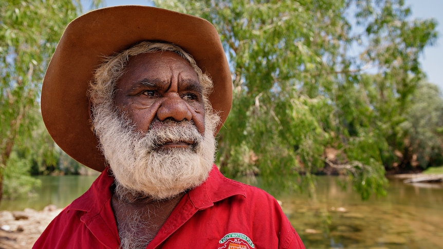 A serious-looking man in a hat an a red shirt looking to the side, with a river and bushland in the background.