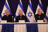 Three men wearing black sitting side by side on a table. A blue and white Israeli flag is in the background.