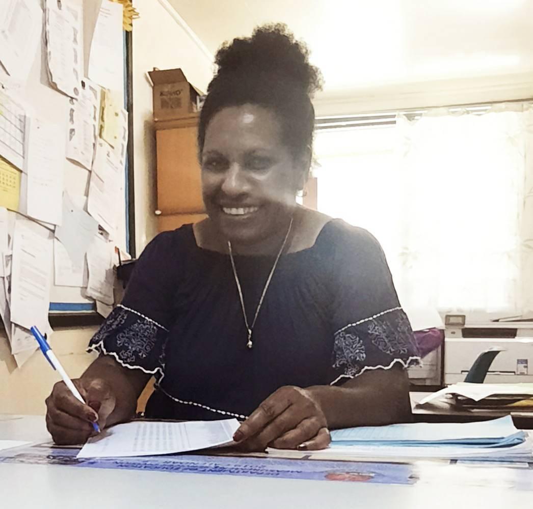 A woman smiling as she sits at a desk with a pen in her hand. 