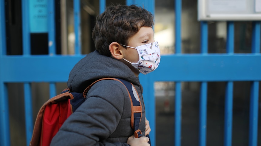 Young boy in winter coat wears face mask in front of blue gates.