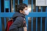 Young boy in winter coat wears face mask in front of blue gates.