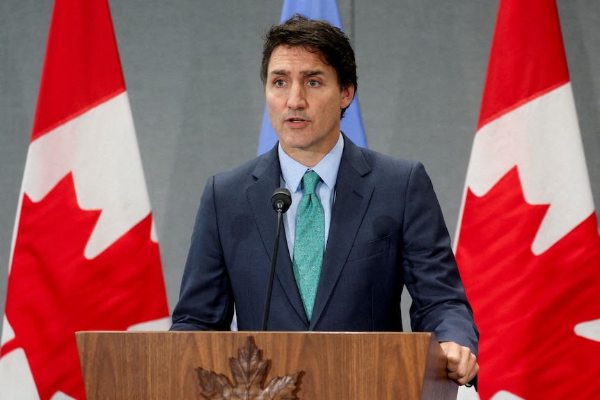 A middle-aged white man with dark hair in a suit stands behind a lectern in front of two red and white Canadian flags.
