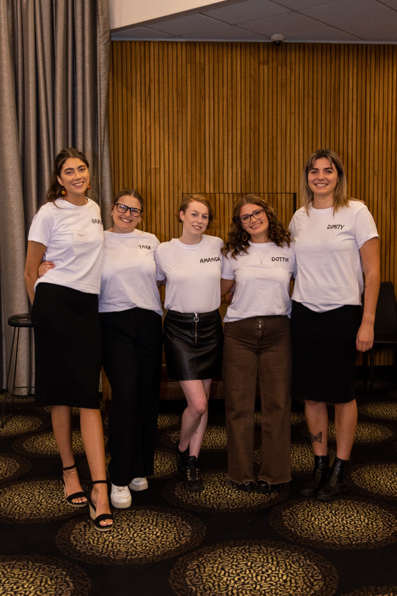 Five young women wearing white T-shirts with a Dare to Dream logo smile, linked arm in arm.