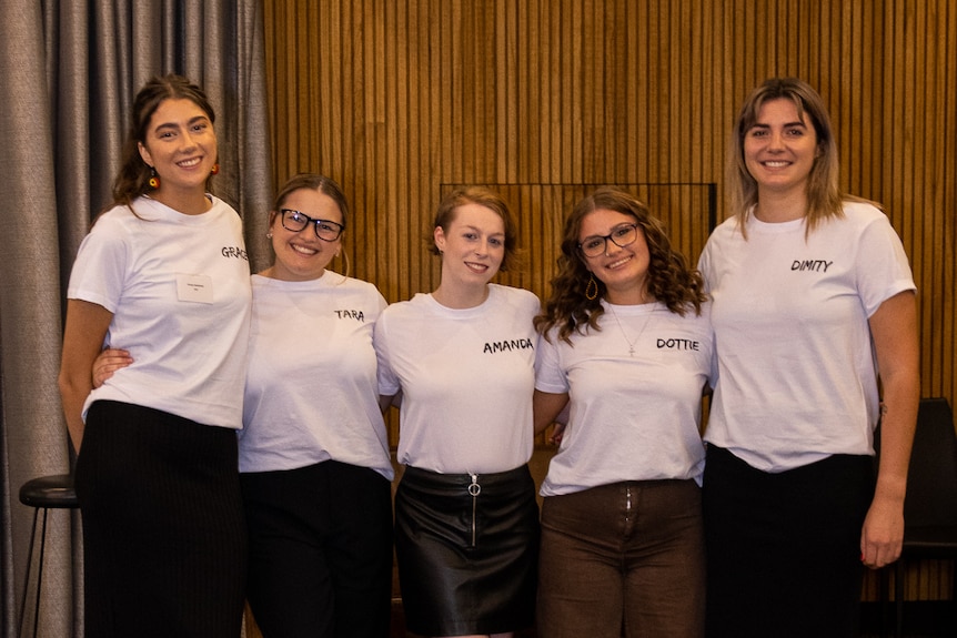 Five young women wearing white T-shirts with a Dare to Dream logo smile, linked arm in arm.