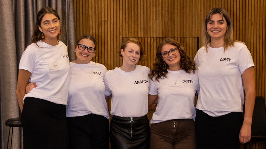 Five young women wearing white T-shirts with a Dare to Dream logo smile, linked arm in arm.