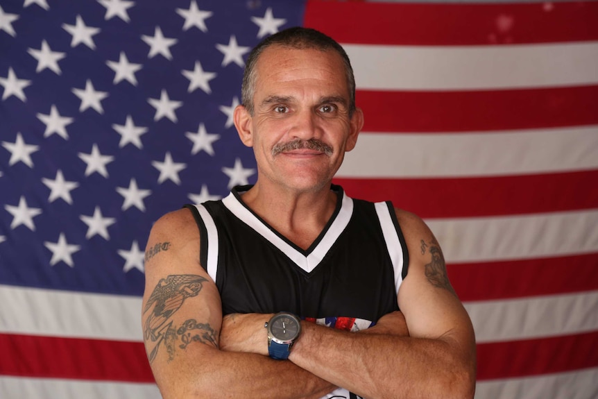 Tattooed man wearing singlet stands in front of US flag.