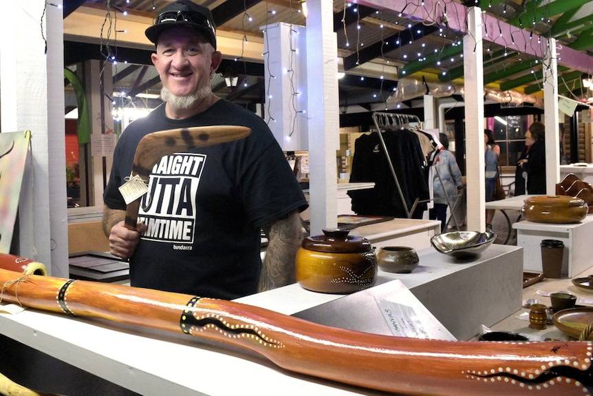 A man stands behind the counter of a market stall holding a boomerang. A polished didgeridoo lies on the counter.