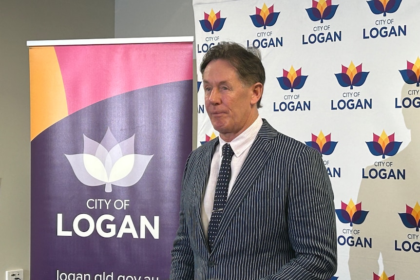 A man wearing a suit standing in front of a purple sign with Logan written on it and a white banner with Logan council branding.