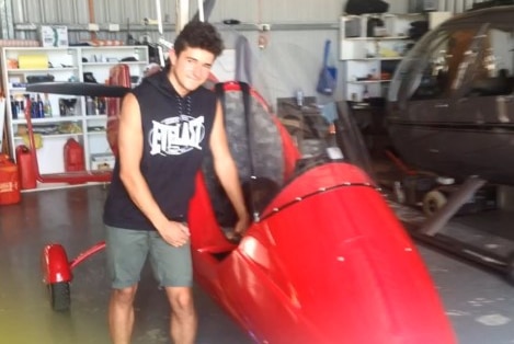 A young man in a black singlet stands next to a red gyrocopter in a hanger.