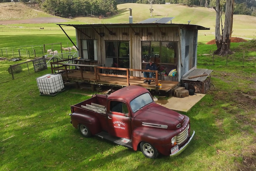 Red pick-up truck parked outside rustic bush shack, a man sits on the deck outside.
