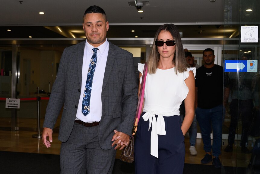 A tall man in a suit walks holding hands with a woman wearing sunglasses and a white top