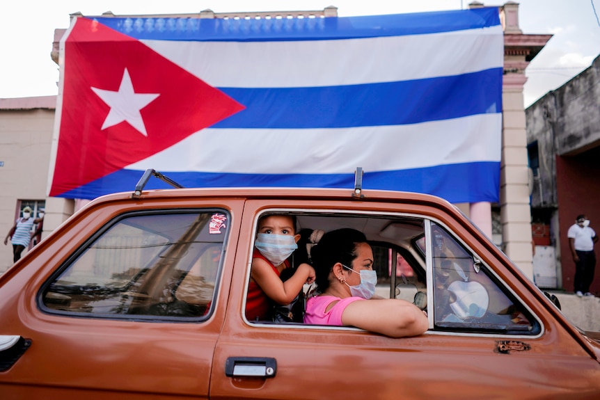 A small boy looks outside a car driven by a woman as they roll past a Cuban flag