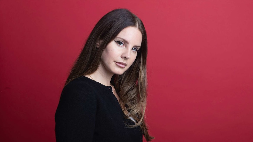 Singer Lana Del Rey poses for a portrait against a red wall. She wears a black cardigan.