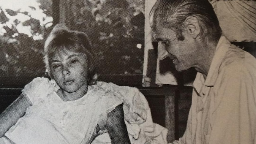 A black and white photo of a teenaged girl reclining on a bed, while a man sits next to her