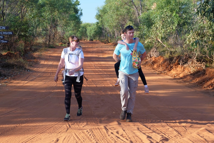 A man and woman walking down red dirt track with baby and small child on their backs