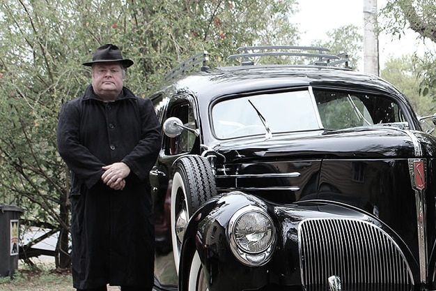Jack Sims wearing a black coat and a black hat next to a black car.