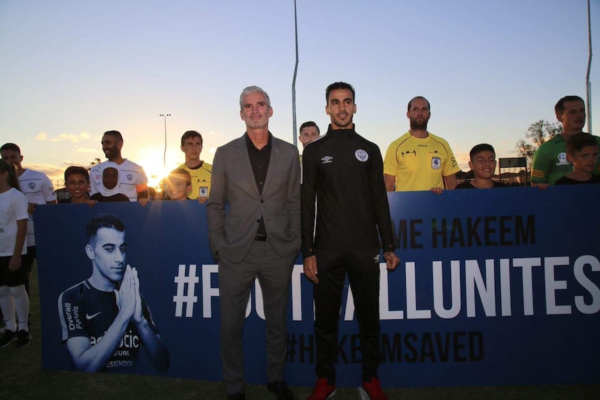 Hakeem and Craig Foster stand in front of a sign reading '#footballunites #hakeemsaved'.