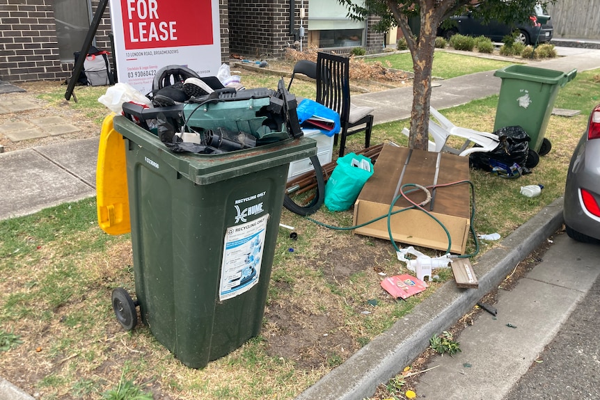 A recycle bin overflows with electrical and other waste while furniture and bags litter the grass footpath.