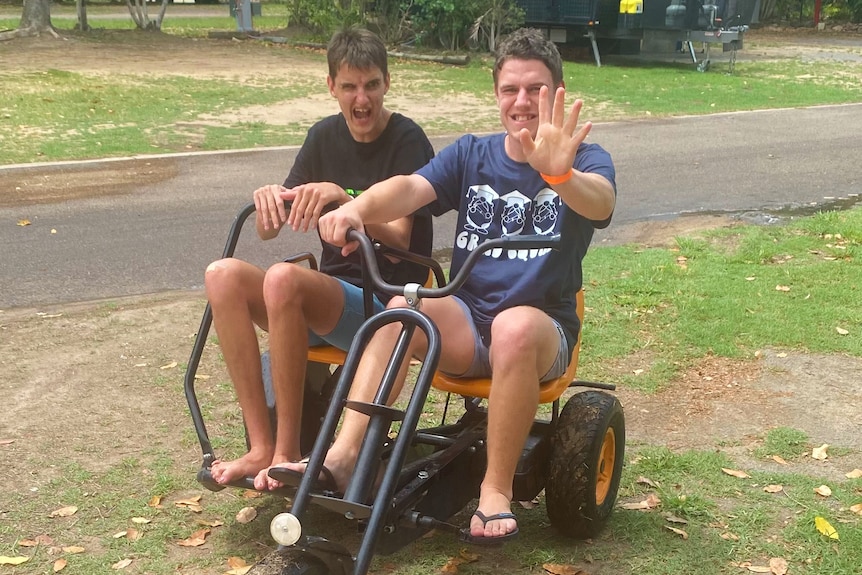 Two men riding a bike, one waves at the camera.