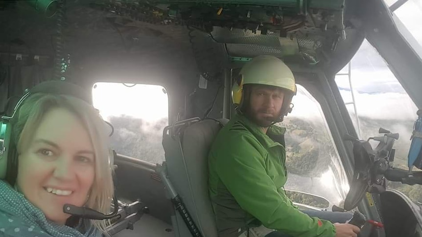 A smiling woman with blonde hair sits in a helicopter beside the pilot, a bearded man wearing a helmet.