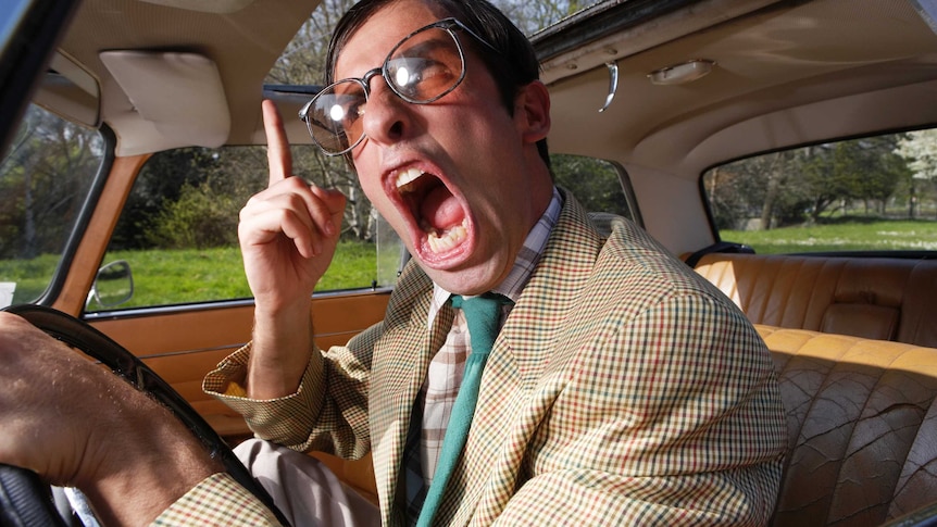 An open mouthed man gesticulating driving an old car wearing a check jacket and glasses