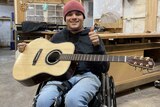 A smiling young man in a wheelchair holds a guitar and gives a thumbs up.