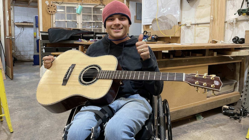 A smiling young man in a wheelchair holds a guitar and gives a thumbs up.