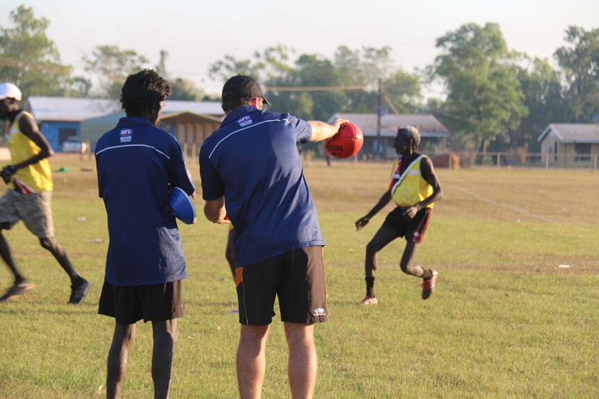 Two men in blue AFL shirts on a football field with their backs to the camera, one with a football in his outstretched hand.