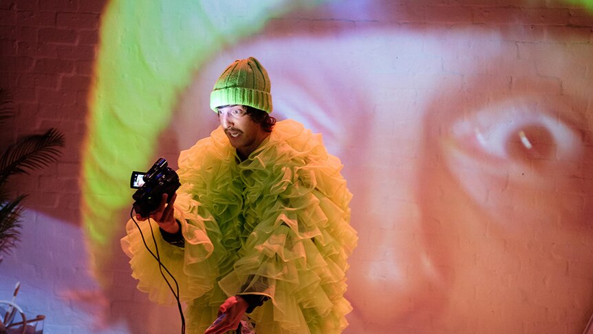 A man with fluro beanie and ruffle tulle jacket points video camera with torch towards own face, which is projected behind him.