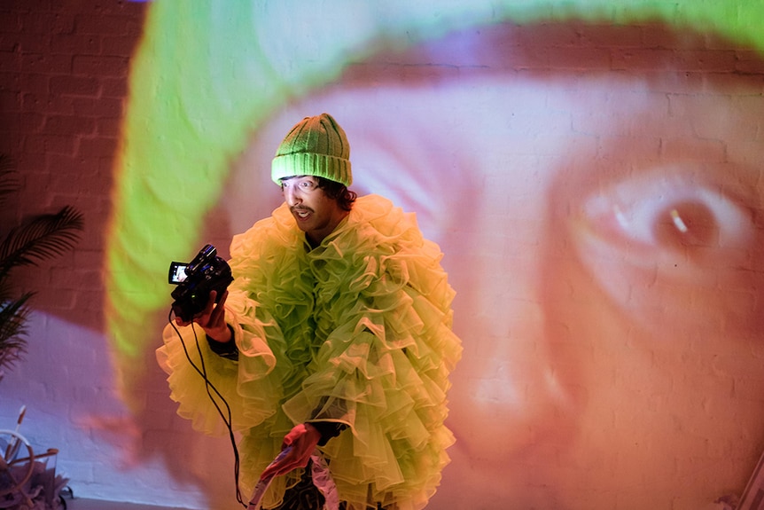 A man with fluro beanie and ruffle tulle jacket points video camera with torch towards own face, which is projected behind him.