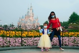 A small girl, wearing a face mask, waves in front of Shanghai Disneyland, supported by her mother.