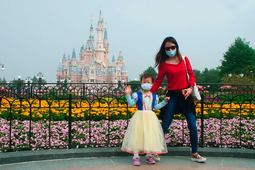 A small girl, wearing a face mask, waves in front of Shanghai Disneyland, supported by her mother.