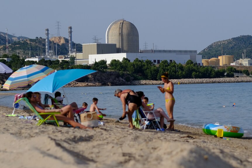 Beachgoers enjoy the sun at a peaceful-looking waterfront, while a nuclear power plant sits in the background.