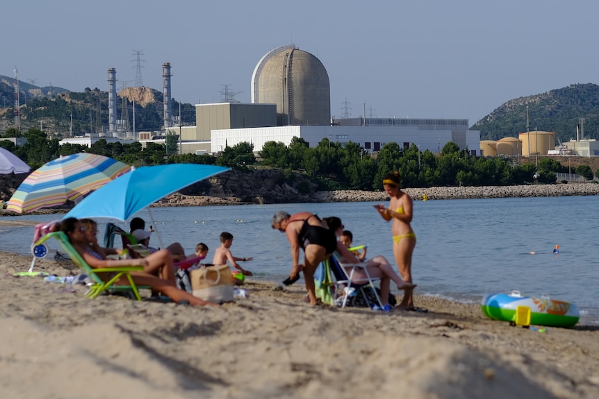Los bañistas disfrutan del sol en un paseo marítimo de apariencia pacífica, mientras una planta de energía nuclear se encuentra en el fondo.