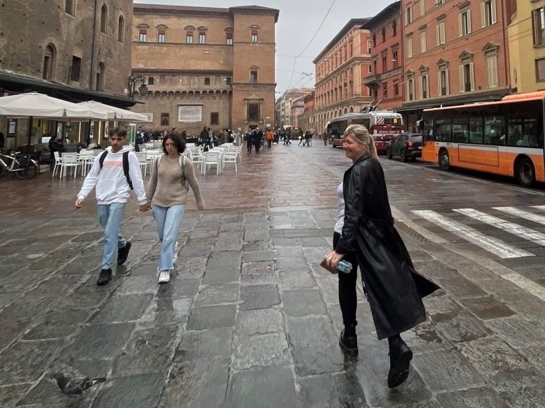 A woman with a black long coat walking and smiling back at the camera in an Italian city