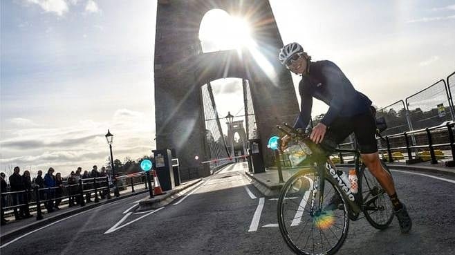 Young man on bike in front of bridge with sun shining through crowd of people lining the side