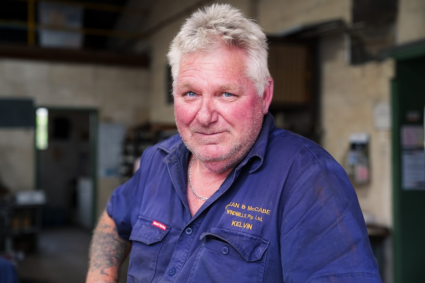 A man with grey hair and blue eyes in a navy shirt stands in a workshop with a hand on his right hip.