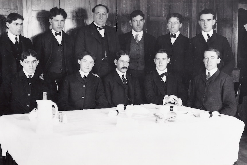 12 well dressed young men stand facing a camera. Black and white image.