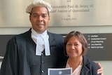 A barrister wearing a white wig in his legal robes stands with his arm around a woman holding a certificate of admission