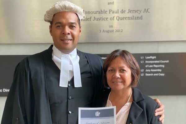 A barrister wearing a white wig in his legal robes stands with his arm around a woman holding a certificate of admission